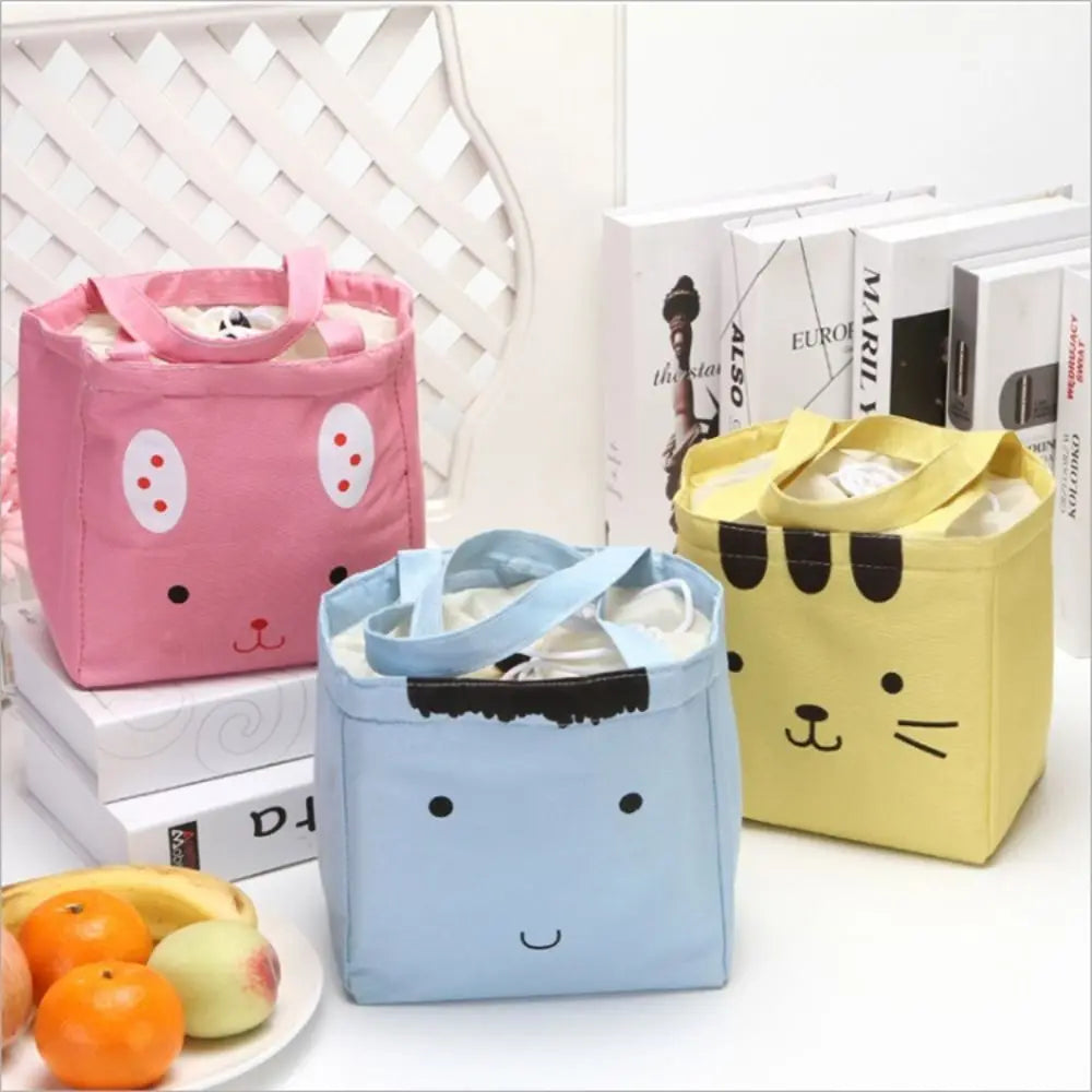 Three cartoon-style kawaii lunch bags in pink, blue, and yellow on a white surface with books and fruits in the background.