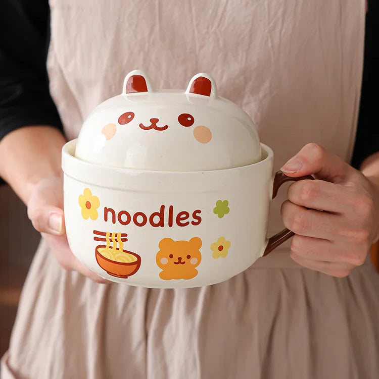 Person holding a cartoon-themed noodle kawaii ramen bowl with a lid, featuring the word 'noodles' and cute animal face design.