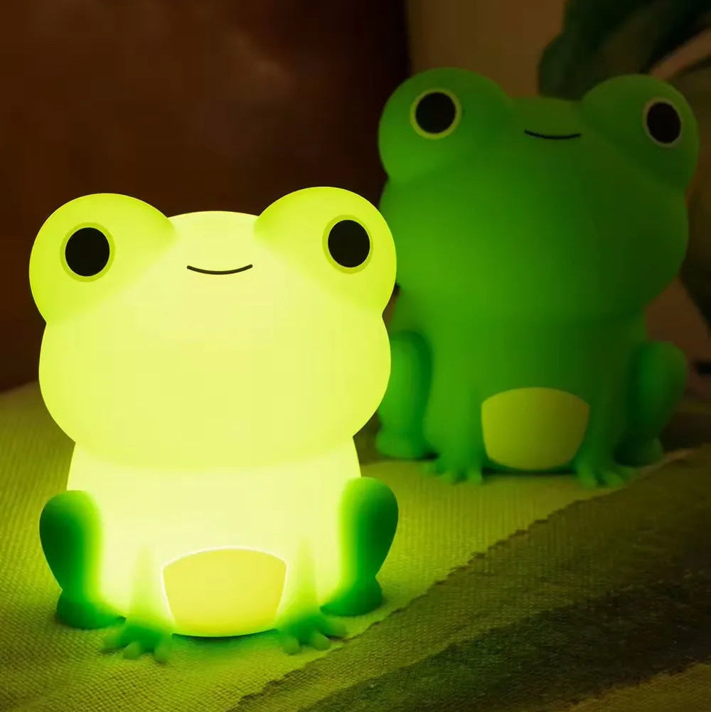 Two frog-shaped kawaii night lights, one glowing green and the other standing, on a soft surface.