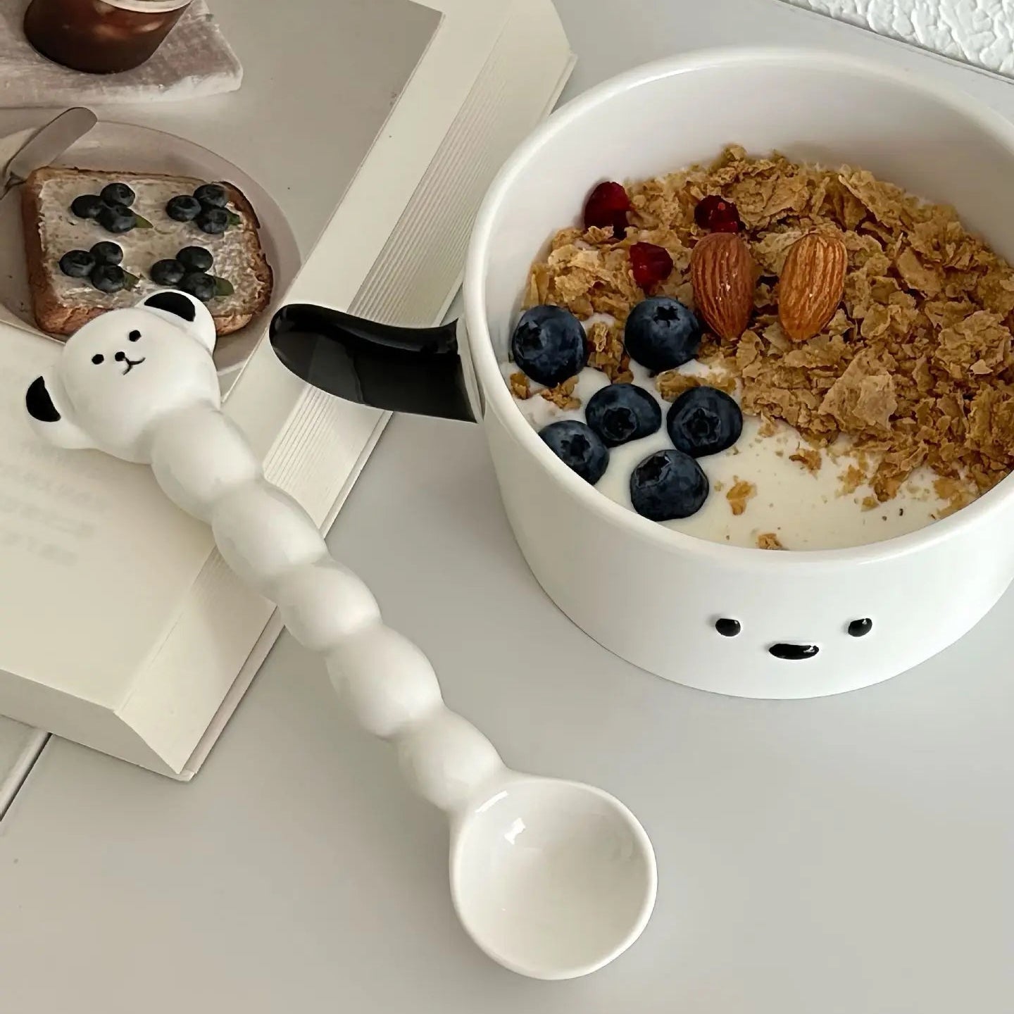 Ceramic kawaii ramen bowl with cereal and fruit, accompanied by a panda-shaped spoon on a table.