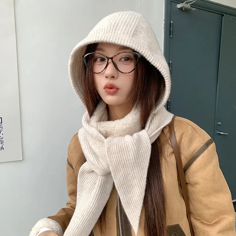 Person wearing a beige coat, white Kawaii knitted hat, and scarf in front of a gray wall.