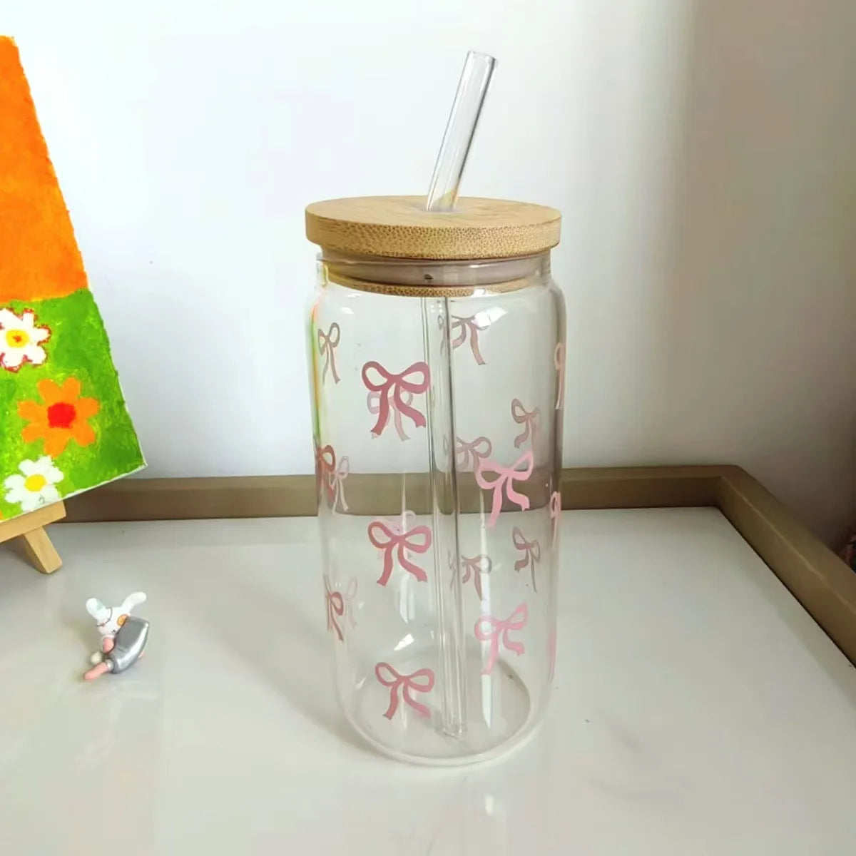 Clear glass kawaii tumbler with wooden lid and pink bows on a white surface.