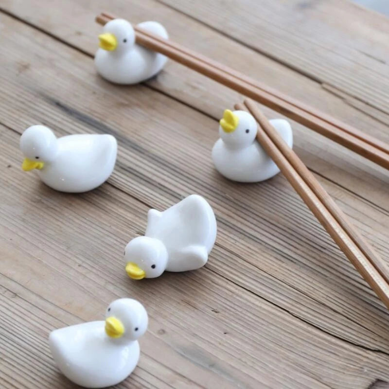 Five white duck-shaped kawaii chopstick holders on a wooden surface with two pairs of wooden chopsticks.