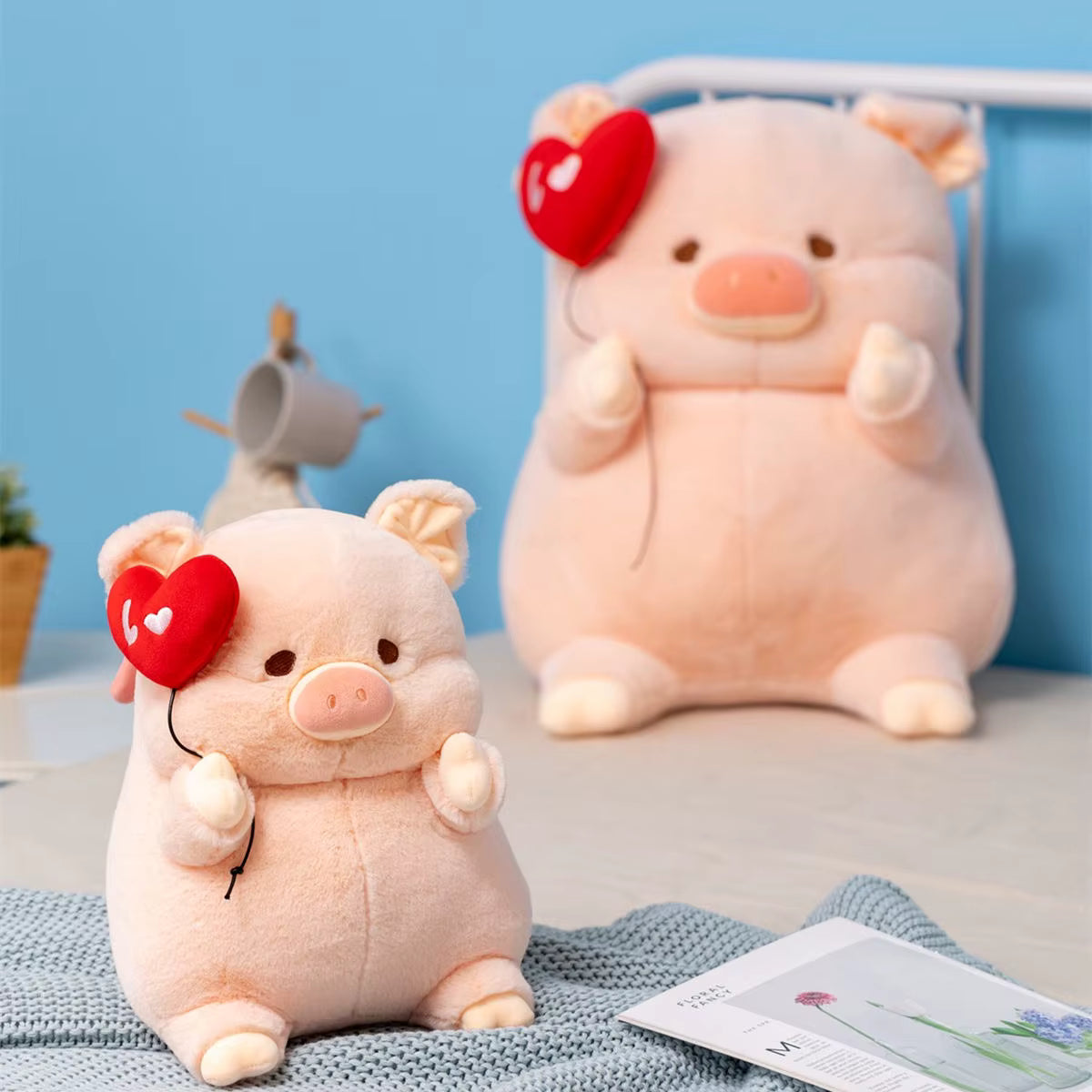Two cute pink plush pig toys holding red heart-shaped balloons, styled on a light blue blanket against a blue wall background.