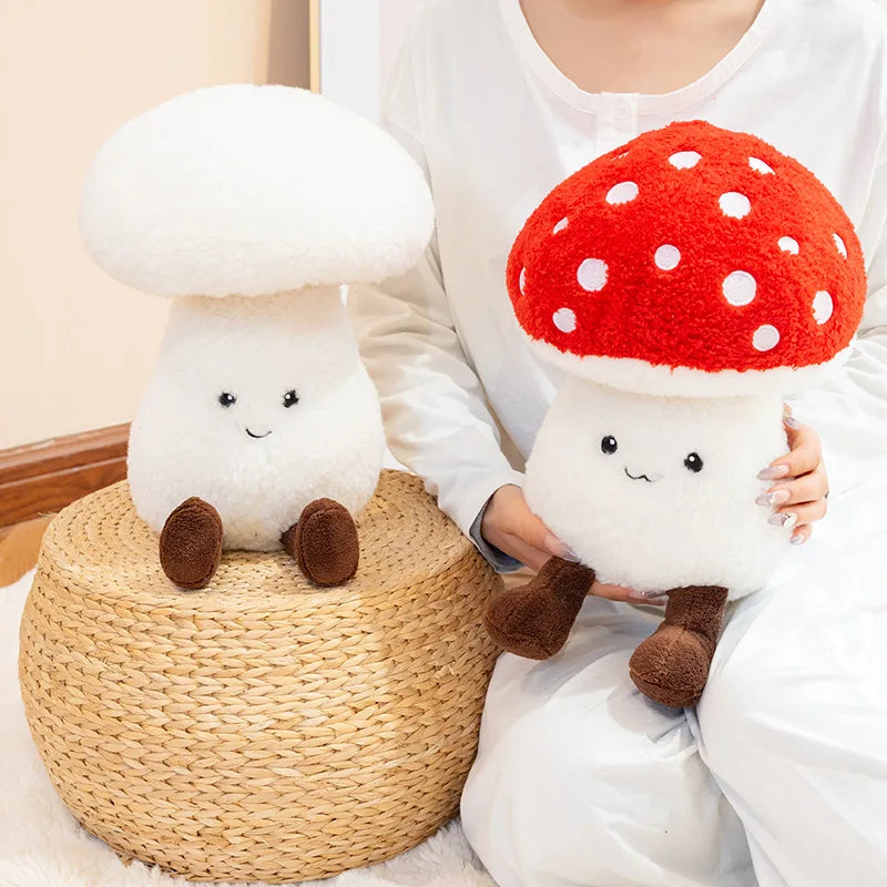 Person holding a kawaii plush mushroom toy with a red cap and white spots, sitting next to another plush mushroom toy on a woven stool.