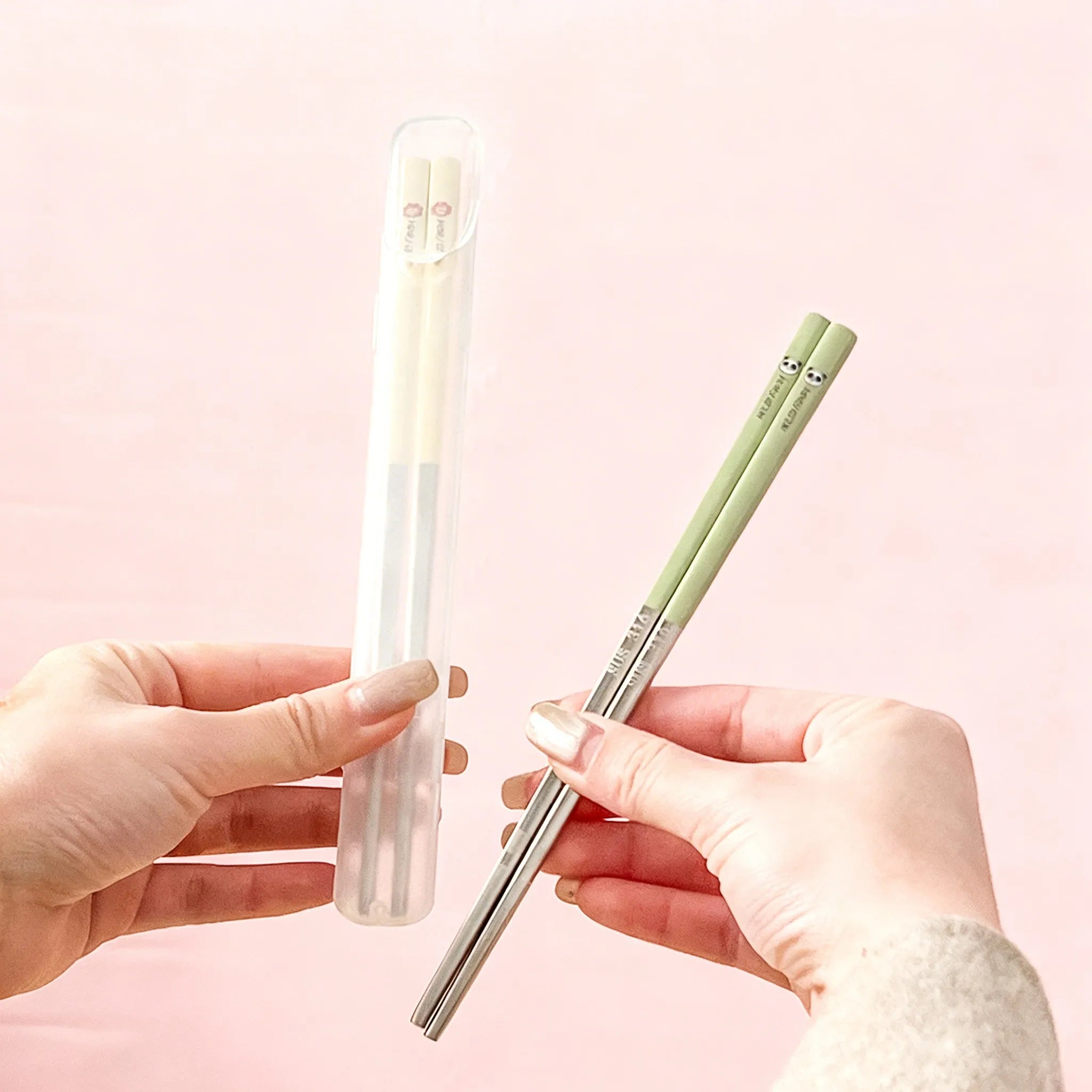 Two hands holding a green kawaii chopsticks and a clear cylindrical case against a pink background