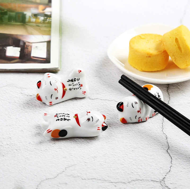 Three kawaii cat-shaped ceramic on a white surface with chopsticks and a plate of food in the background.