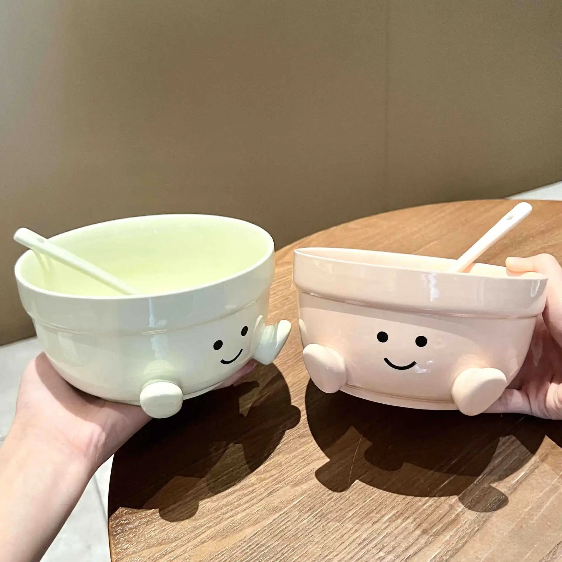 Two cartoon-style ceramic kawaii bowls with faces, arms, and legs on a wooden surface.