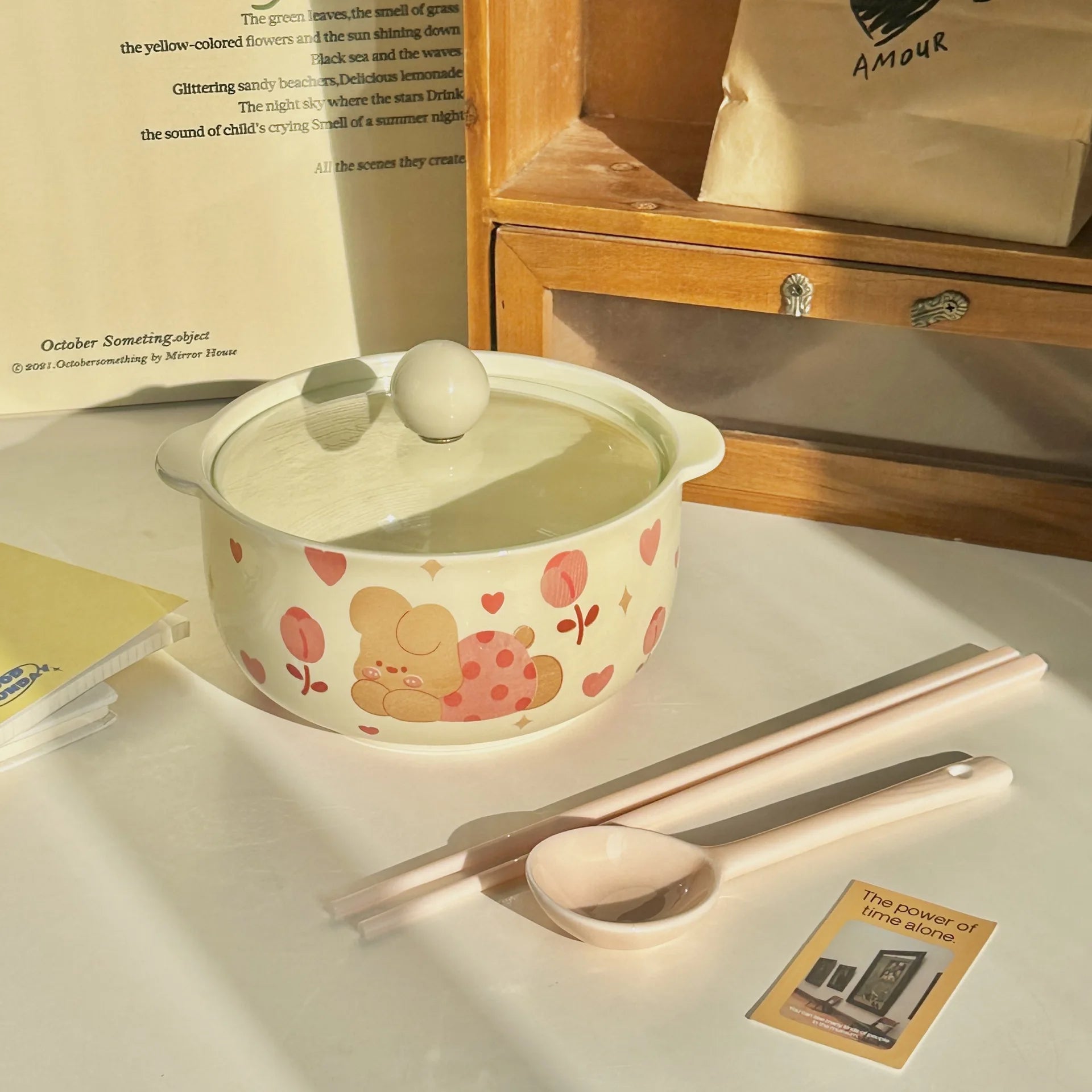 Ceramic kawaii ramen bowls with lid, spoon, and chopsticks on a table with a book in the background