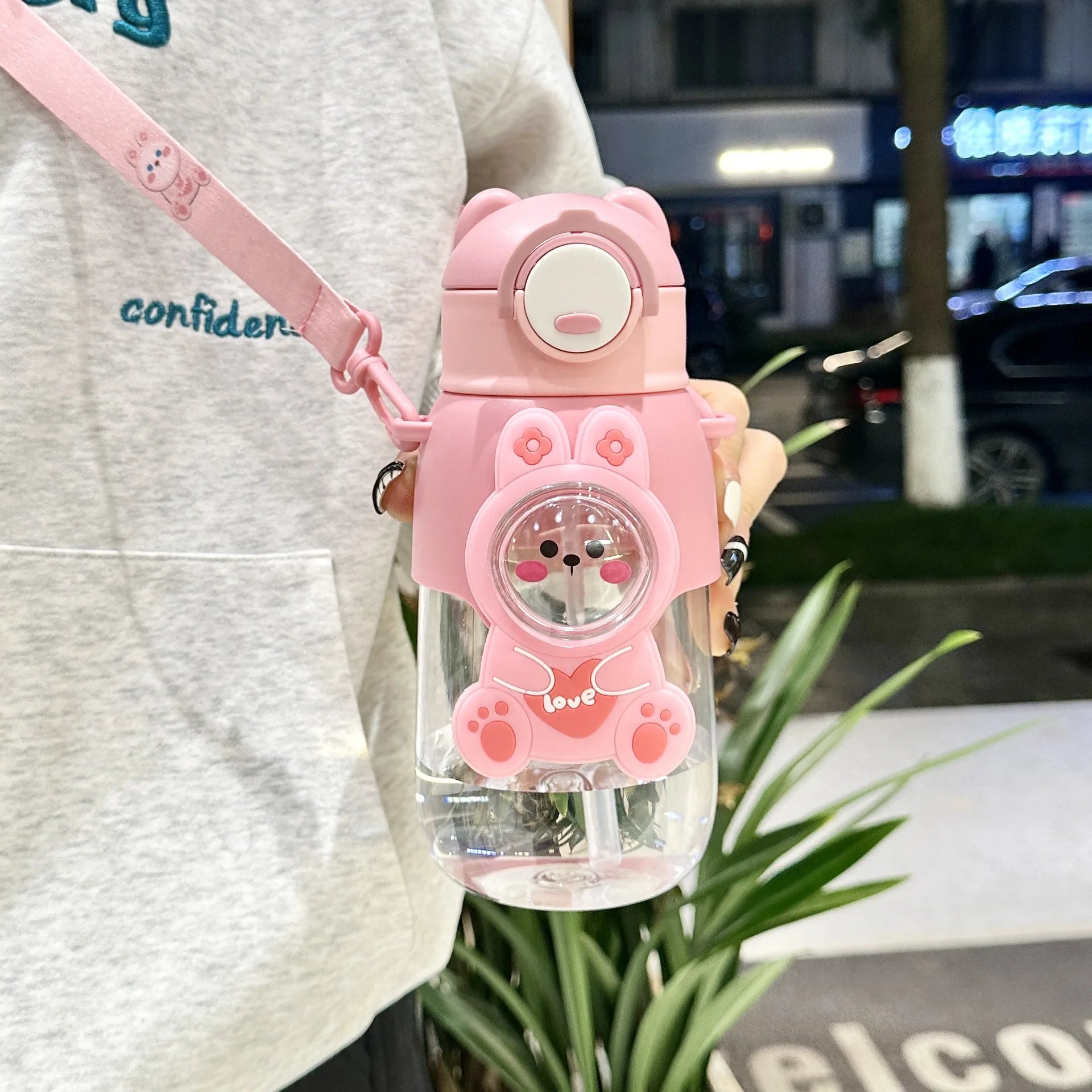 Pink cartoon kawaii water bottle with a clear body held by a person in an urban setting.