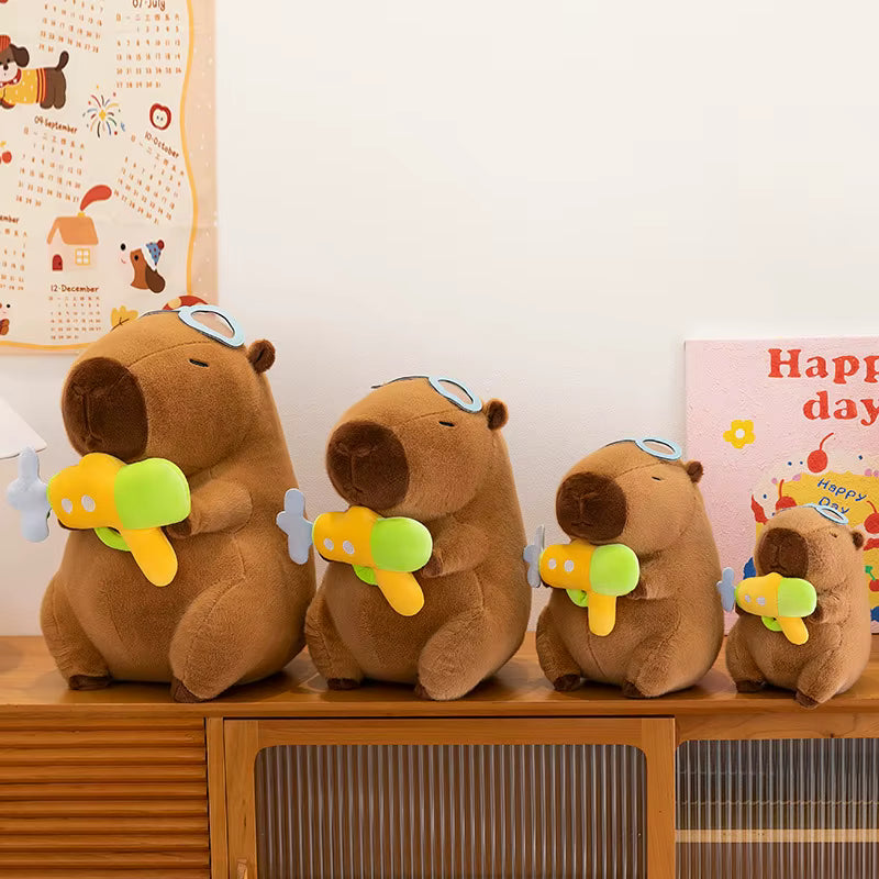 Four brown capybara plush toys of different sizes sitting on a wooden cabinet, each holding a small yellow and green toy airplane. The background shows a white wall with a calendar and a colorful card.