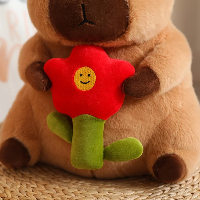 Capybara plush toy holding a red flower with a smiley face on a woven surface.
