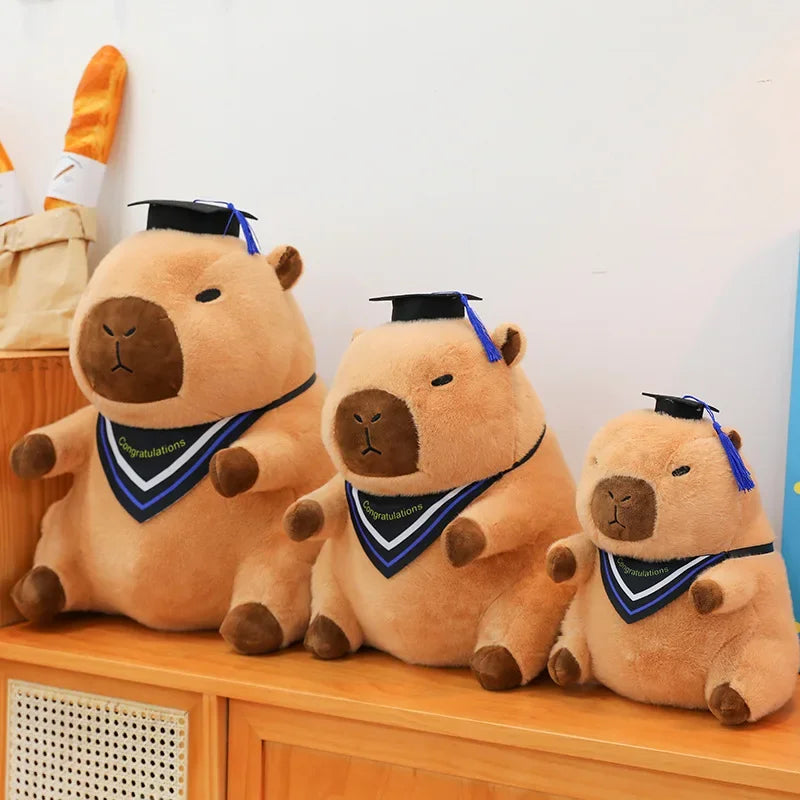 Three kawaii plush toys resembling beavers with graduation caps and sashes on a wooden surface.