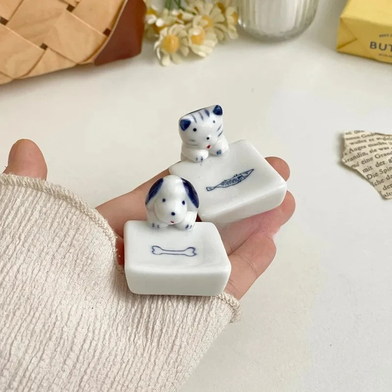 Hand holding two small kawaii chopstick holders dog figurines on a light surface with a woven basket and flowers in the background.