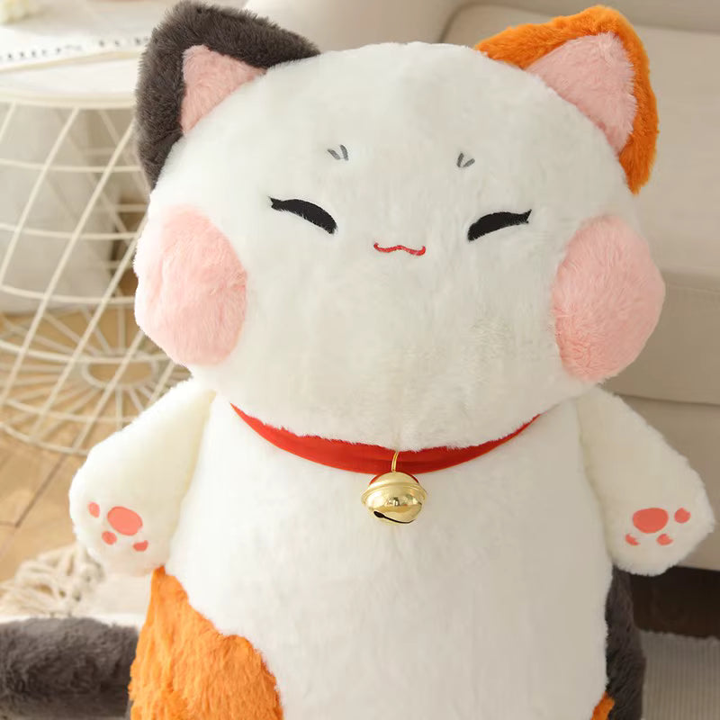 A plush calico cat toy with a white body, orange and dark gray patches, pink inner ears and cheeks, a red collar with a small gold bell, and closed embroidered eyes, photographed indoors against a light neutral background.