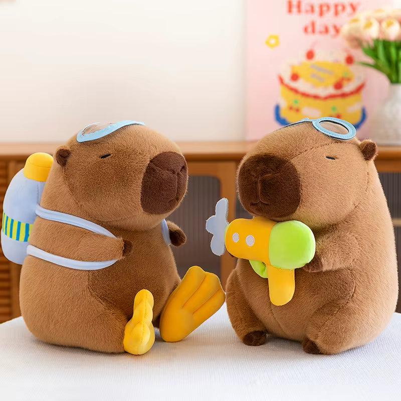 Two brown capybara plush toys sitting side by side on a white surface. One toy wears blue swim goggles and carries a blue and yellow backpack, while the other wears blue swim goggles and holds a yellow and green toy airplane.