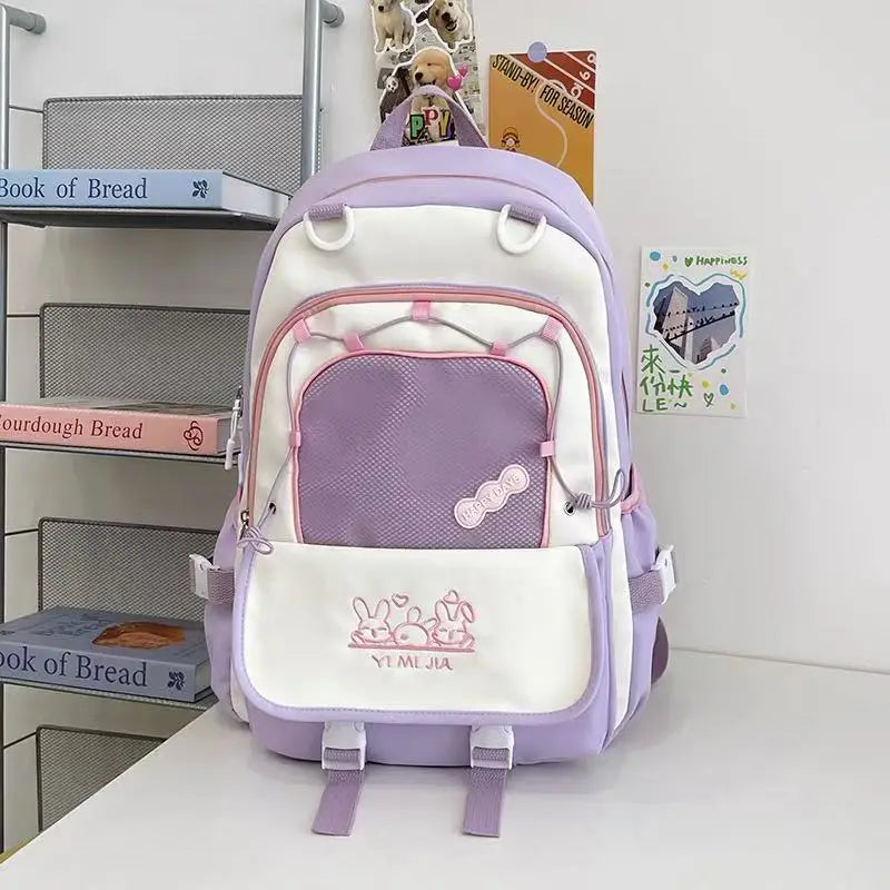 A pastel purple and white school backpack with pink zipper accents and cartoon bunny graphics on the front pocket, displayed on a white table against a white wall with decorative books and cards.