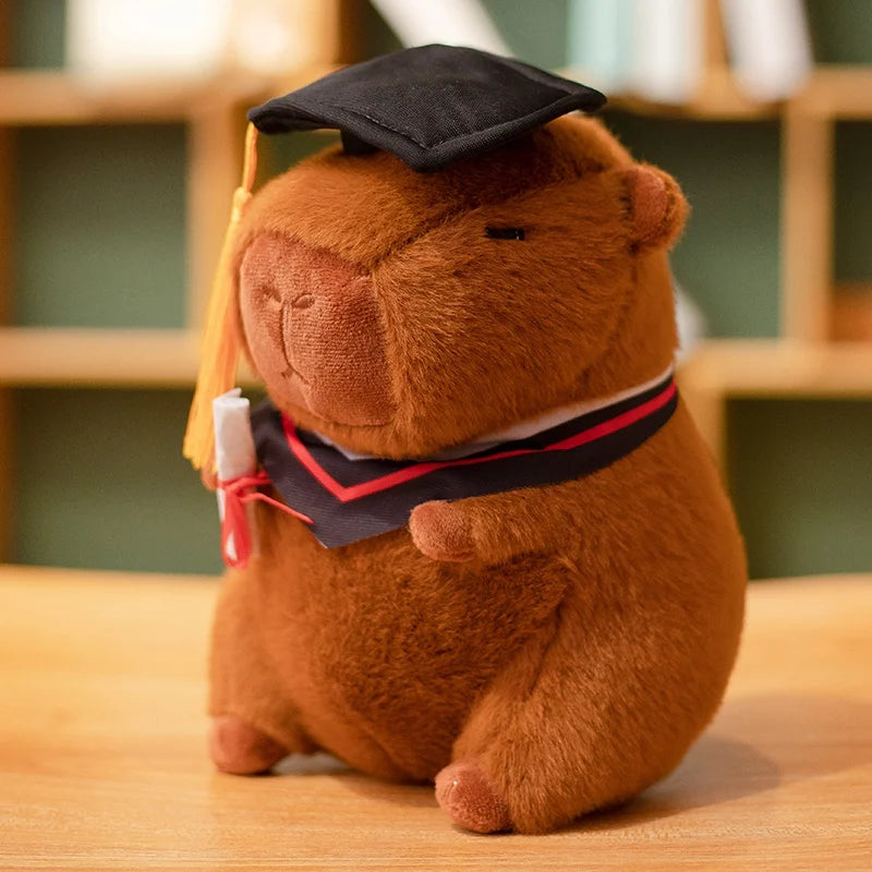 Capybara plush toy resembling a bear wearing a graduation cap and gown on a wooden surface.