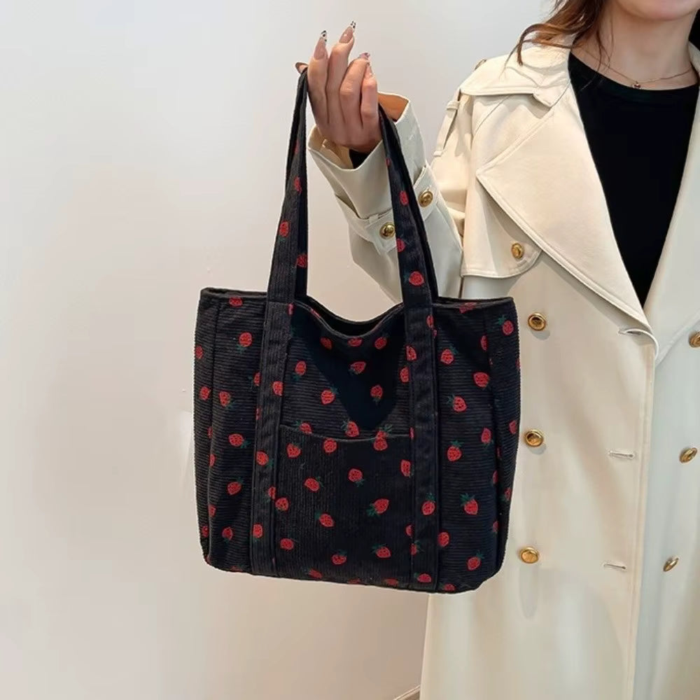 A woman wearing a white coat holds up a large black tote bag with a red strawberry print in an indoor setting.