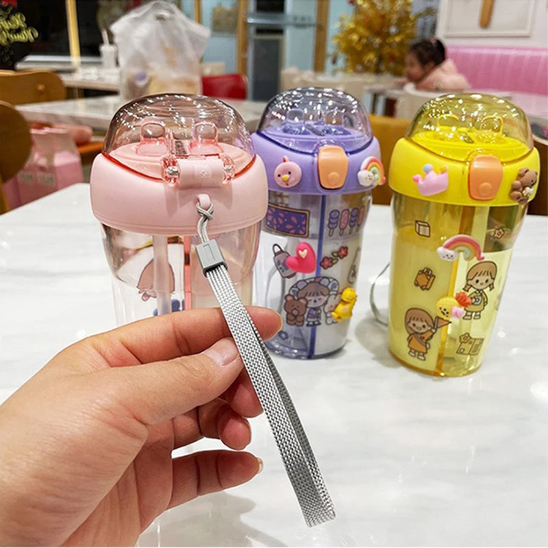 Three colorful children's kawaii cups with cartoon designs on a table, one held by a hand with a metal straw.
