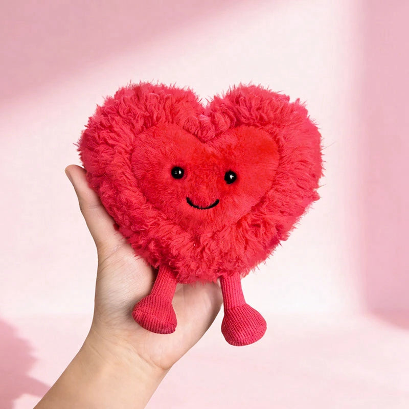 Red heart-shaped kawaii plush toy with a smiling face held in a hand against a pink background