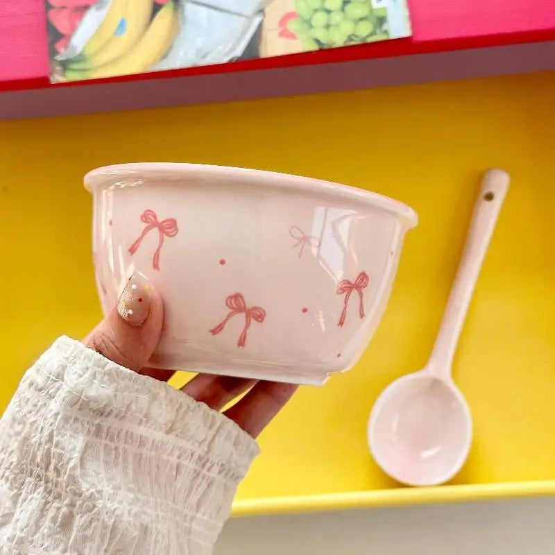 A hand holding a white ceramic bowl decorated with pink bow and dot patterns, with a matching pink spoon resting in a yellow tray behind it.
