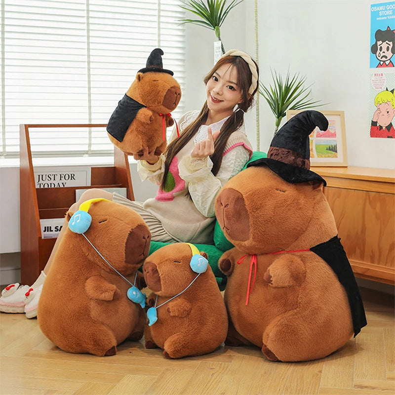 Person sitting on the floor with three large brown capybara plush toys wearing witch hats.