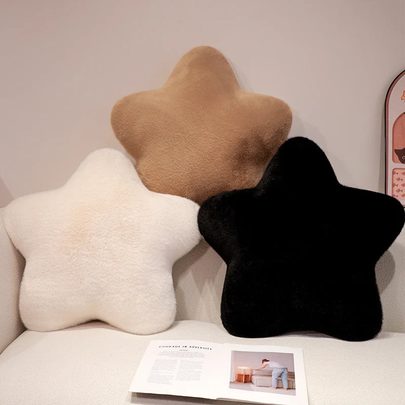 Three plush star-shaped throw pillows in cream, tan, and black, arranged on a light beige sofa with an open magazine in front.