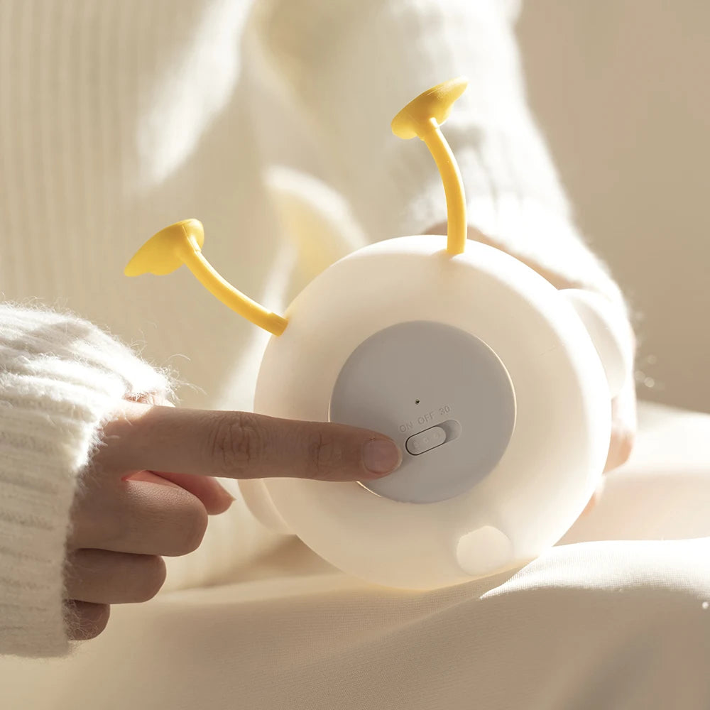 Person holding a round kawaii night light with yellow antenna, pressing a button on a soft beige background.