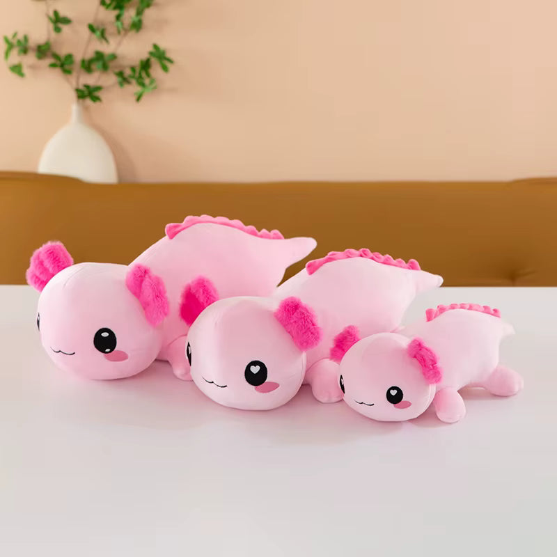 Set of pink axolotl plush toys on a white surface with a beige wall and plant in the background.