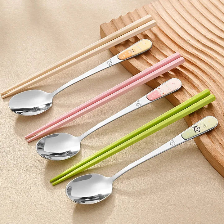 Set of stainless steel kawaii utensils and chopsticks with cartoon character handles on a wooden surface.