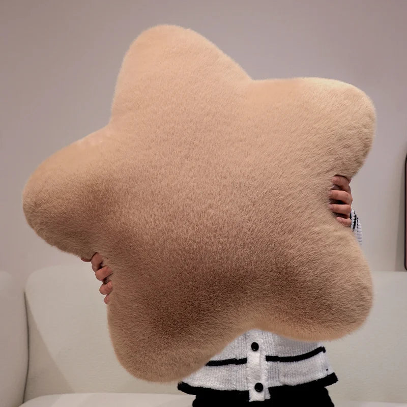 A large plush star-shaped throw pillow in a warm beige color, held by a person sitting on a white sofa.