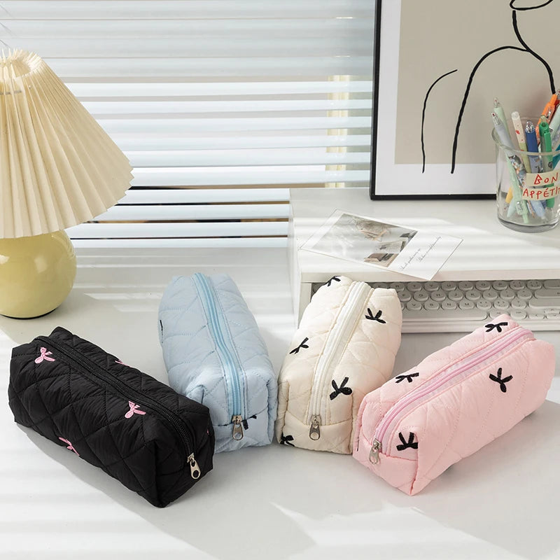 Four quilted kawaii pouches in black, blue, beige, and pink on a desk with a lamp and framed picture in the background.
