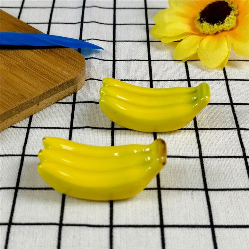Two yellow banana-shaped kawaii chopstick holders on a checkered tablecloth with a wooden board and flower in the background.