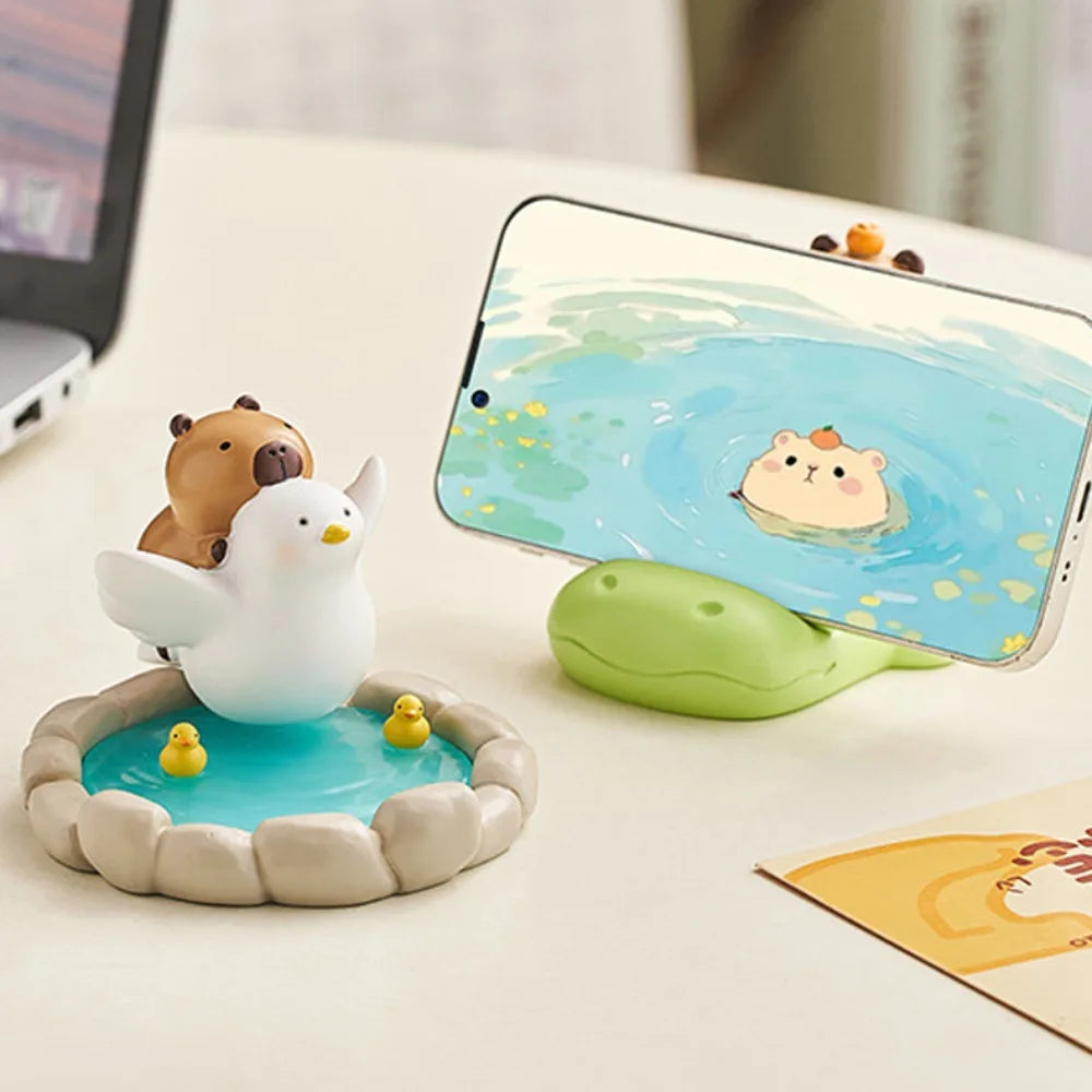 Kawaii phone holder with a small pond figurine, duck, and bear, and a phone displaying a similar scene.