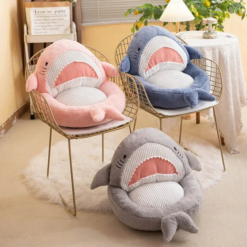 Three shark-shaped kawaii cushions in different colors on chairs in a room.