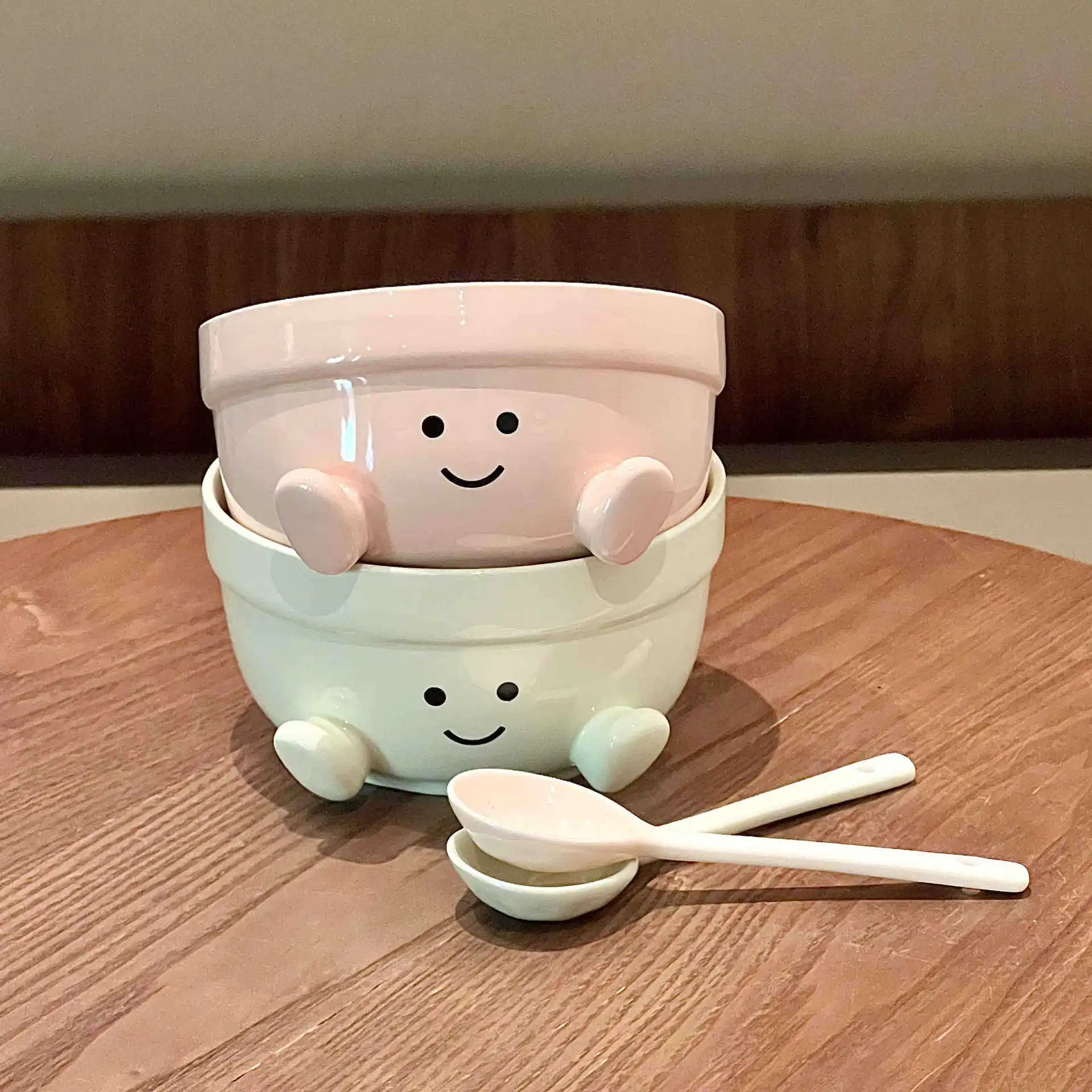 Two stacked ceramic kawaii bowls with cartoon faces and a spoon on a wooden surface