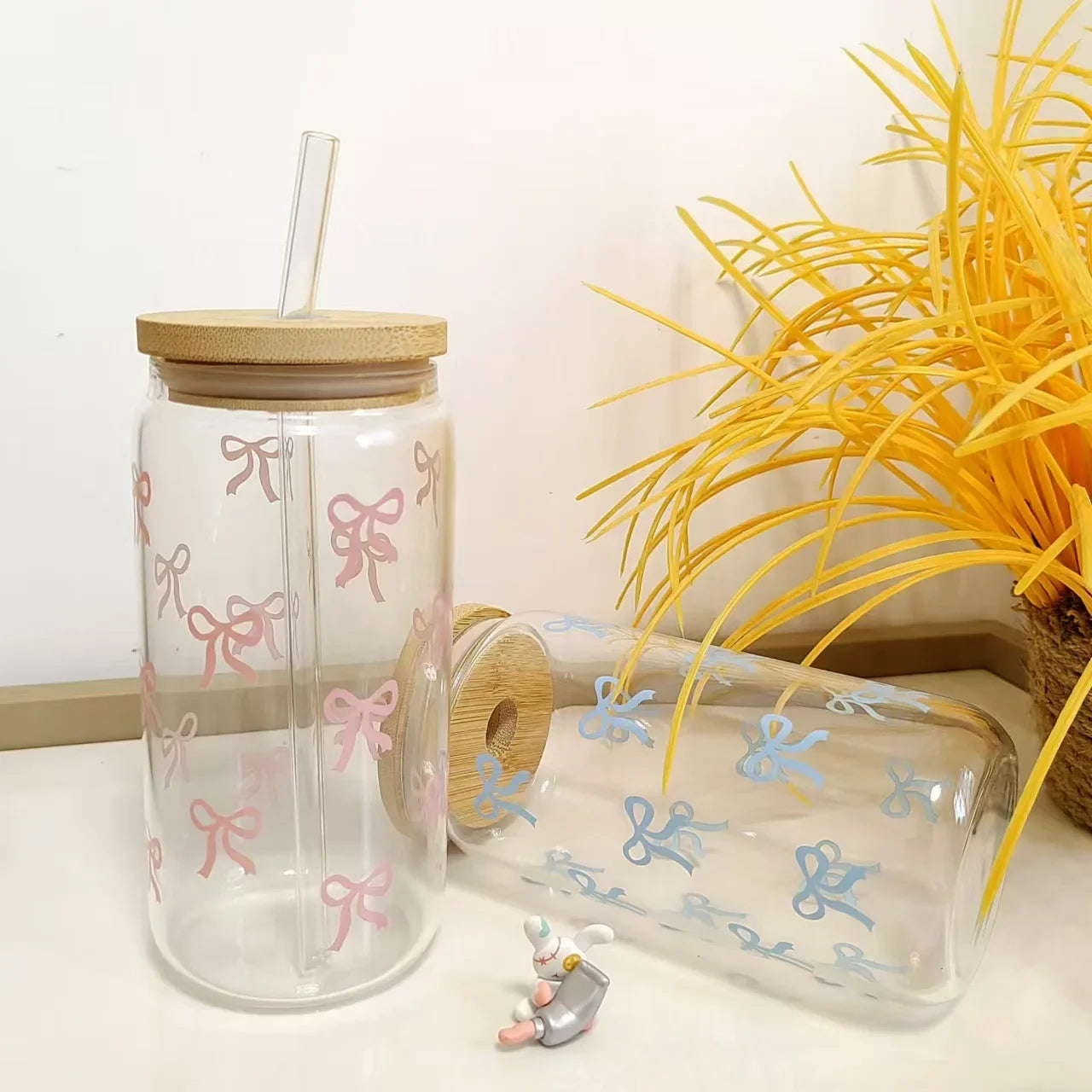Clear glass kawaii tumbler with pink bows and wooden lid on a white surface with yellow decorative elements.