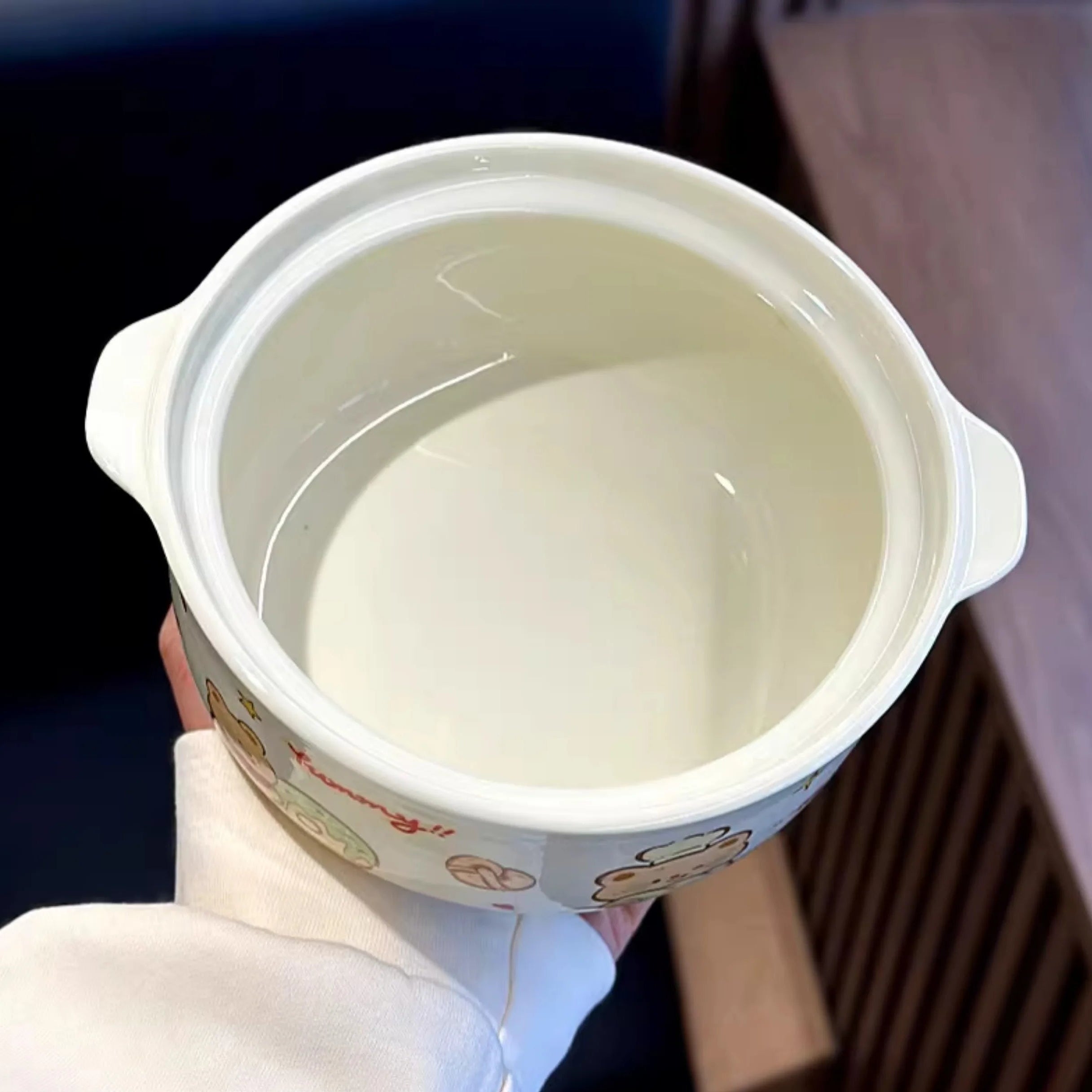 Hand holding a small white kawaii ramen bowl with a lid, filled with a liquid, against a blurred background.
