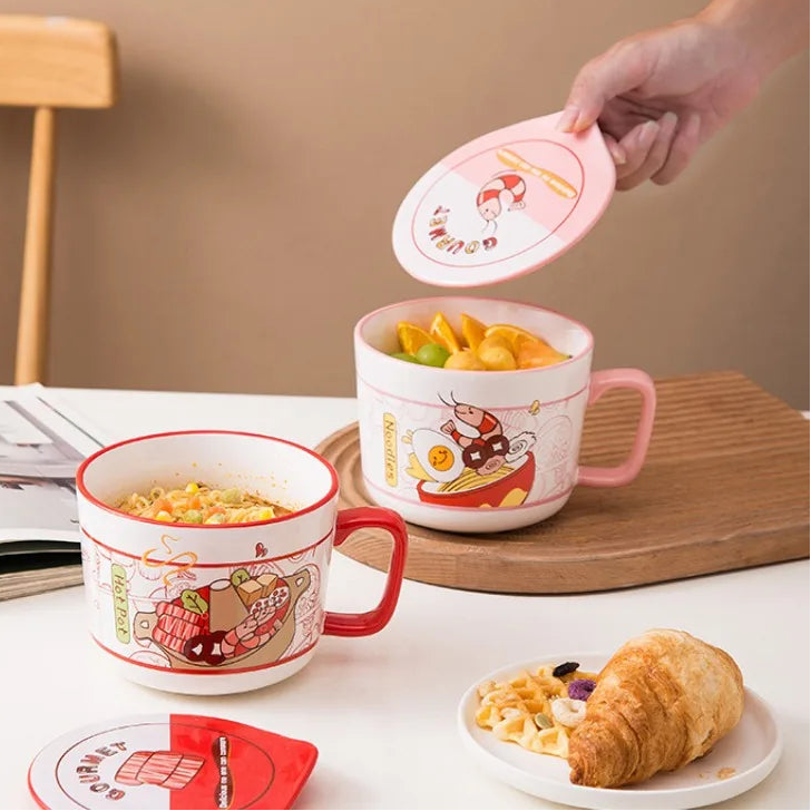 Two ceramic kawaii ramen bowls with cartoon designs on a table with food and a chair in the background.