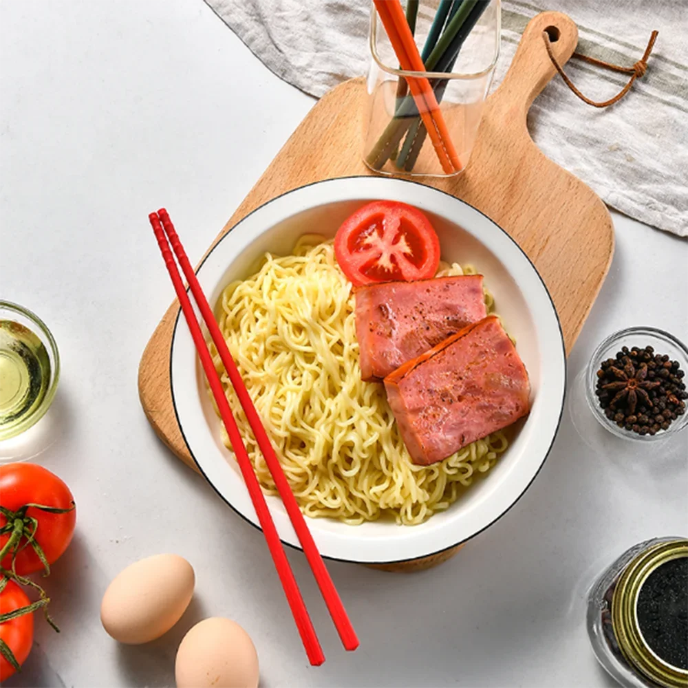 Noodles with sliced ham and tomatoes on a white plate with red kawaii chopsticks, surrounded by ingredients on a light gray background.