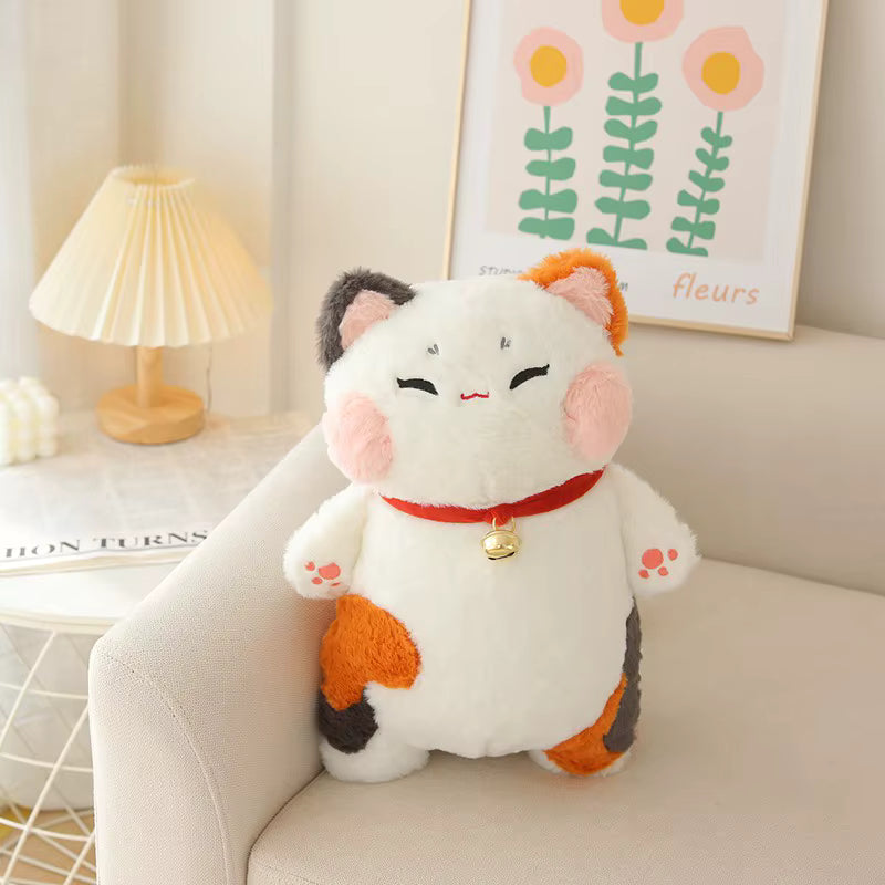 A white calico cat plush toy with orange, black, and pink patches, wearing a red collar with a gold bell, sitting on a beige armchair in a softly lit room.