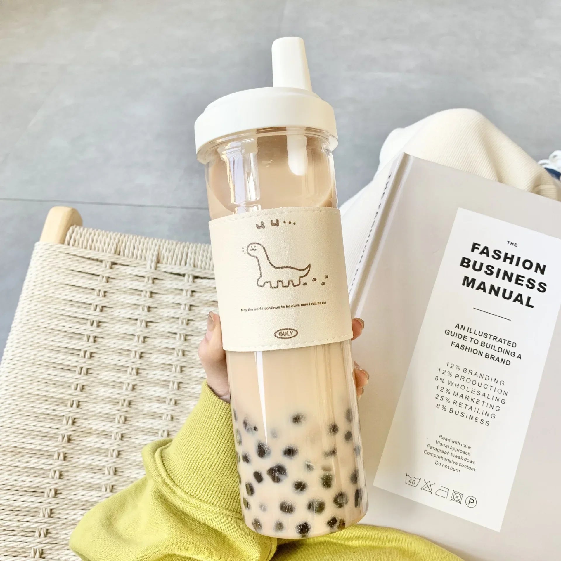 Clear bubble tea kawaii water bottle with a straw and a book titled 'The Fashion Business Manual' on a light gray background.