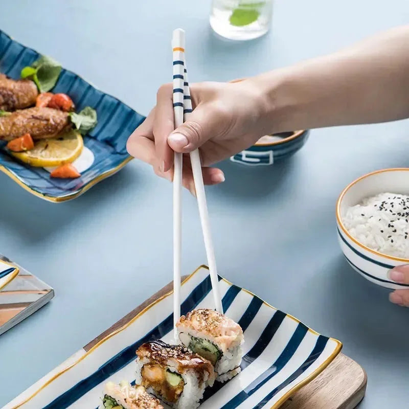 Person using kawaii chopsticks to pick up sushi from a striped plate on a light blue table.