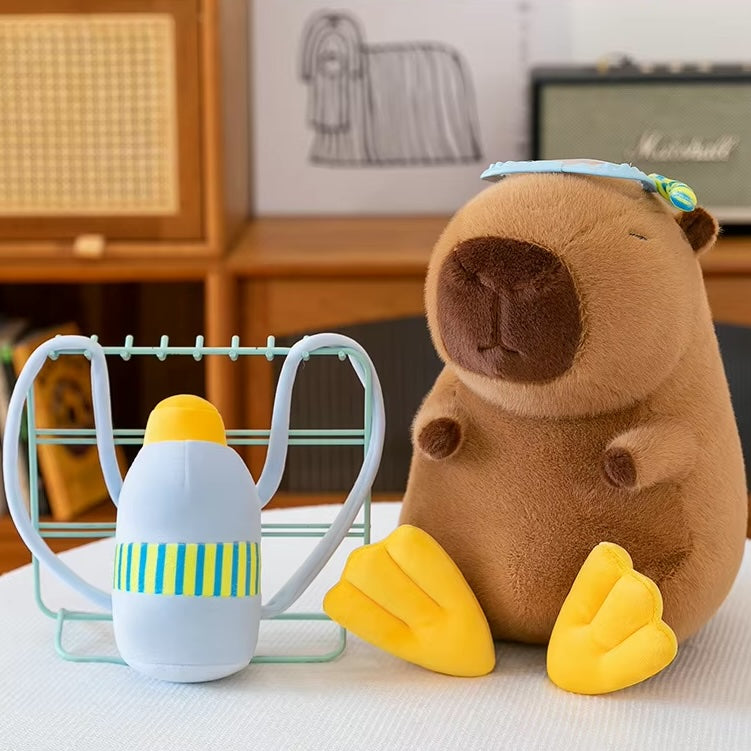 A plush capybara toy sits on a white surface in a styled indoor setting, next to a small white bottle with yellow and blue-green stripes inside a mint green wire holder. The capybara has a brown face and belly, yellow webbed feet, and a light blue headband with a small multicolor accent.
