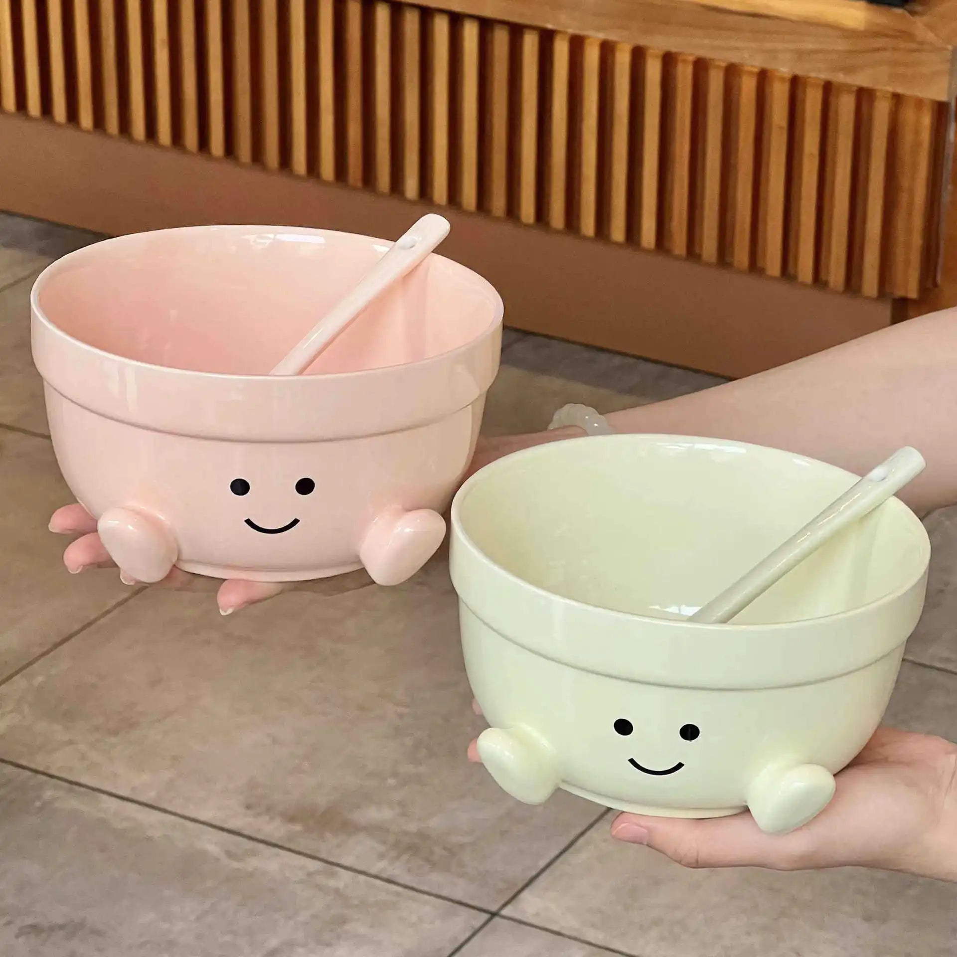Two cartoon-style kawaii bowls with faces, one pink and one green, on a tiled floor.