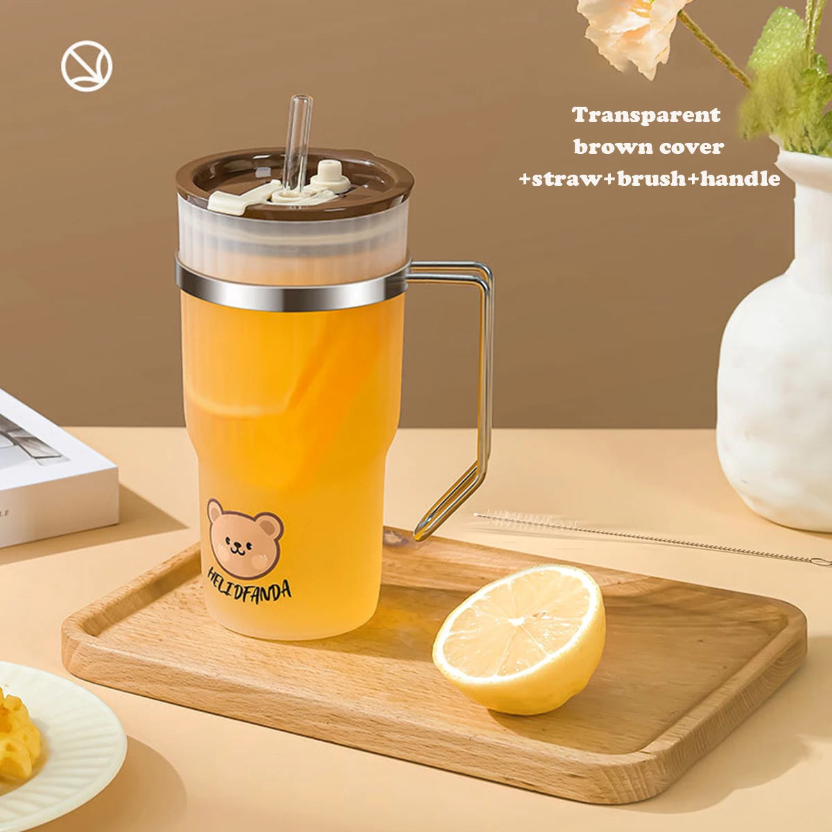 Clear brown cover with straw and handle on a kawaii water cup of orange juice, placed on a wooden tray with a lemon and a vase in the background.