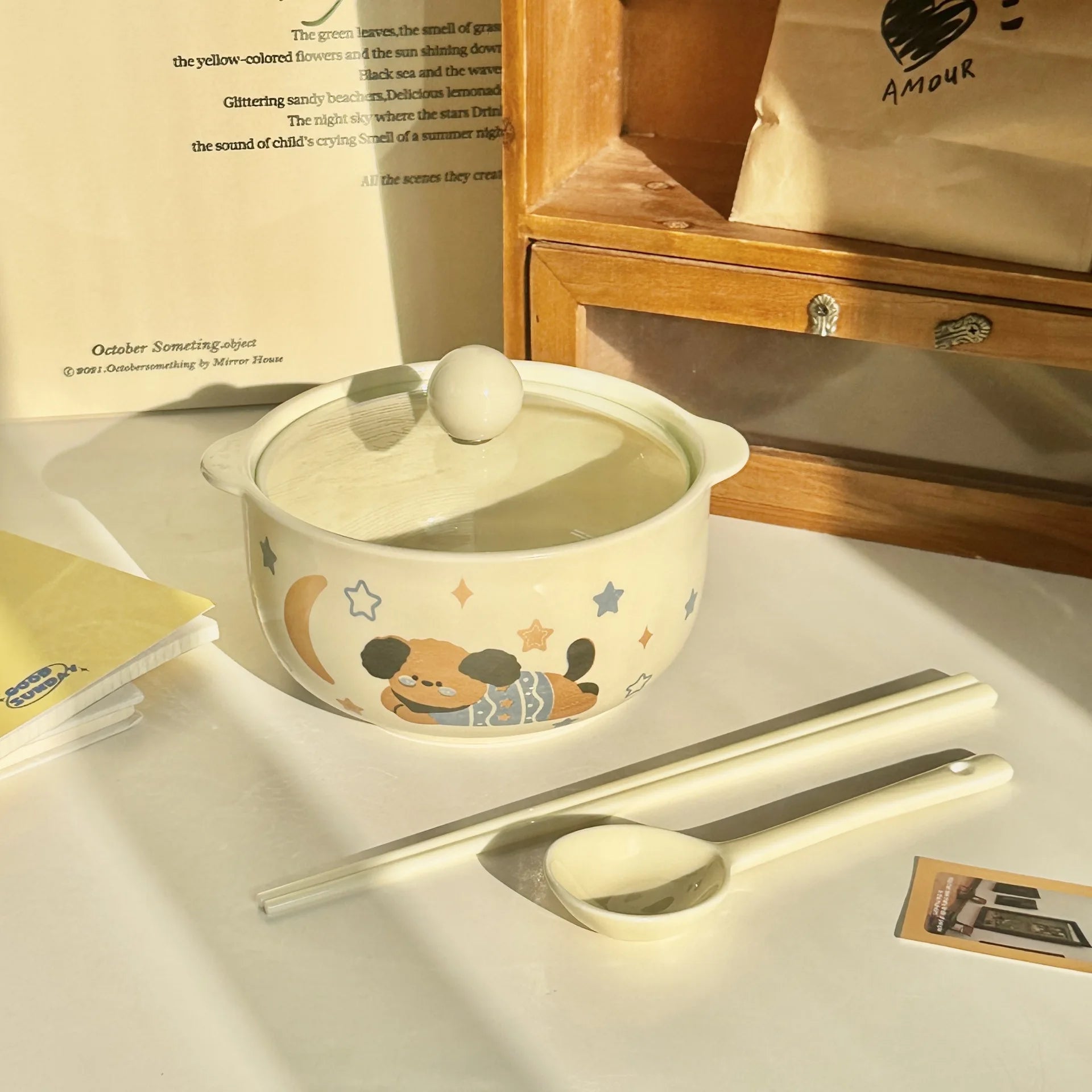 Ceramic kawaii ramen bowl with lid, spoon, and chopsticks on a light surface with a wooden cabinet in the background.