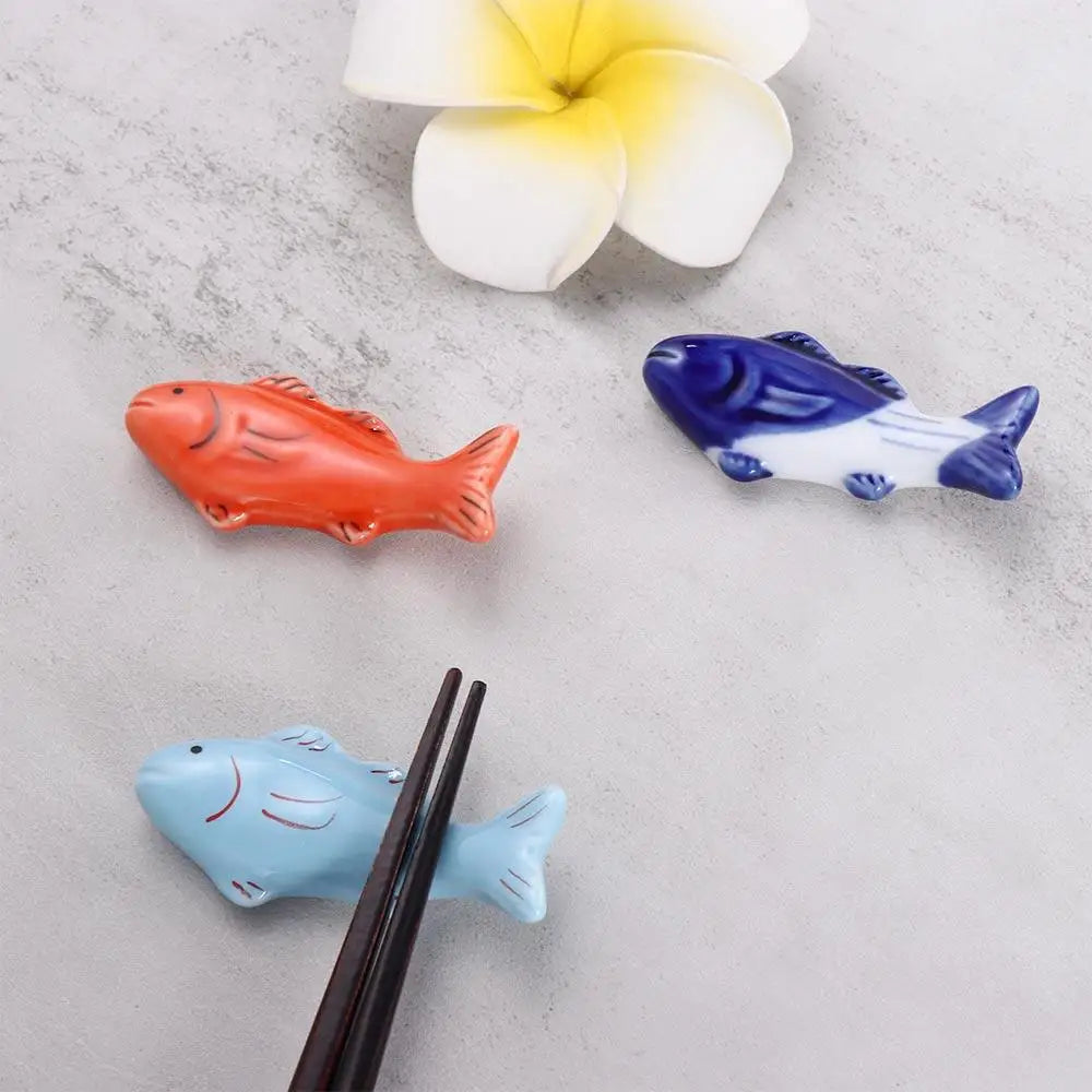 Three fish-shaped ceramic kawaii chopstick rests on a light gray surface with white and yellow flowers.