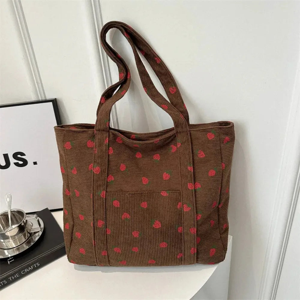 A brown corduroy tote bag with a red strawberry pattern, displayed on a white table next to a book and a coffee cup against a white paneled wall.